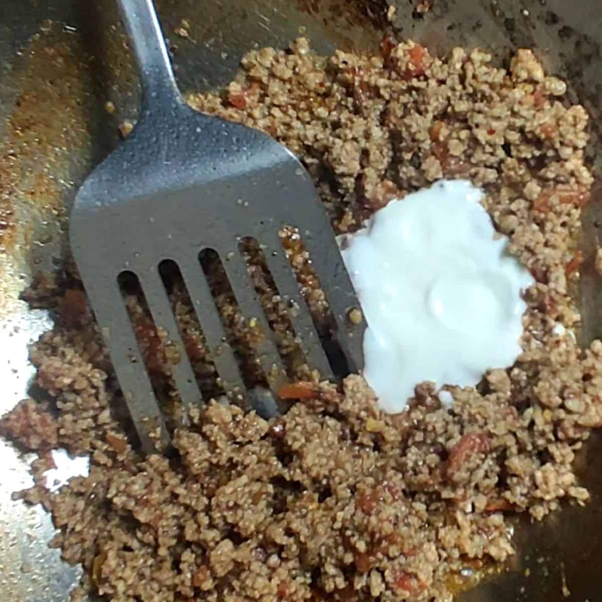 a hand with a spoon pouring fresh yoghurt in beef mince that is cooked in silver wok