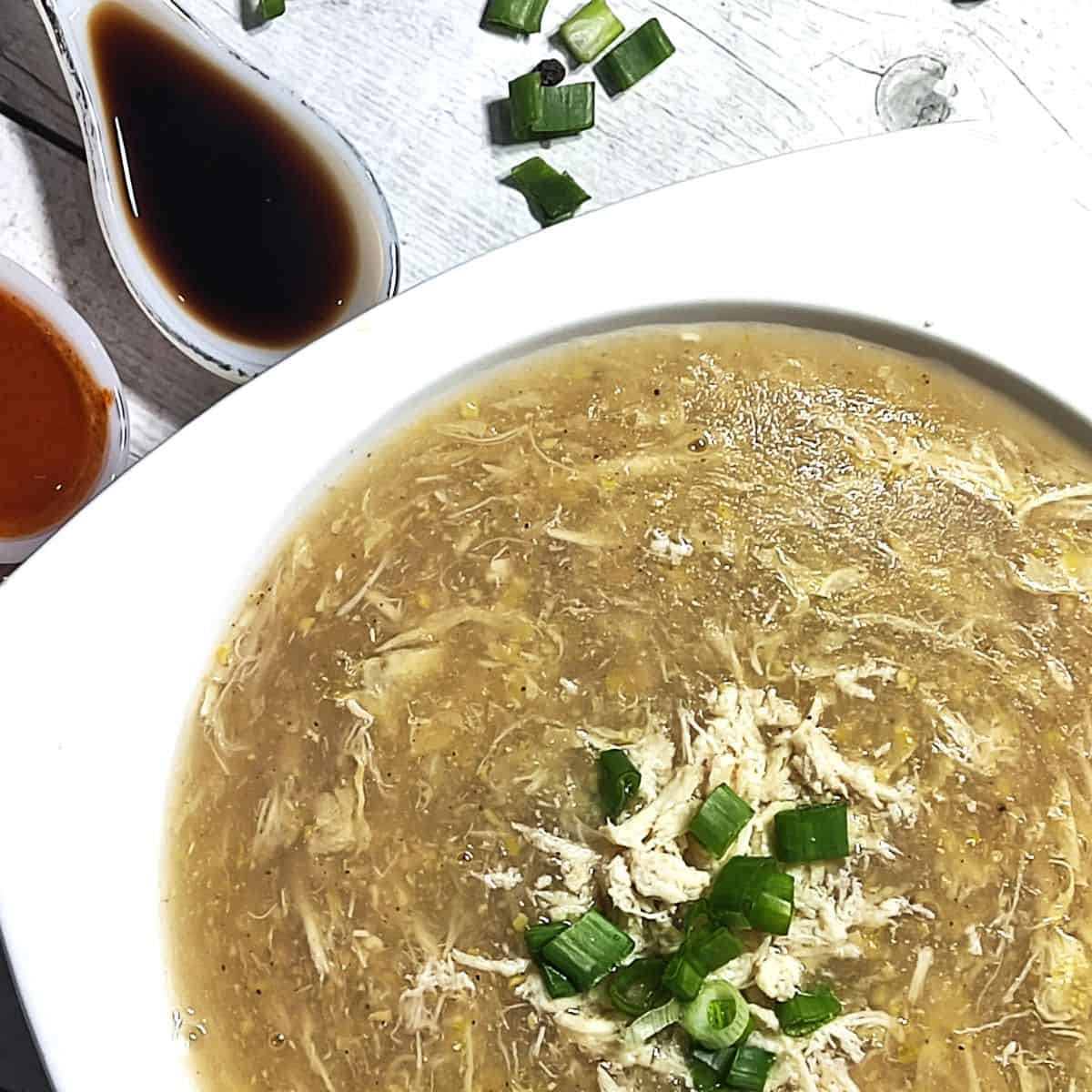 chicken corn soup served in white bowl with spoon on side showing soya sauce and red chilli sauce