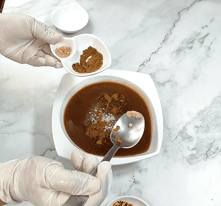 Tamarind chutney being prepared in white bowl with spices and measuring spoon on marble surface.