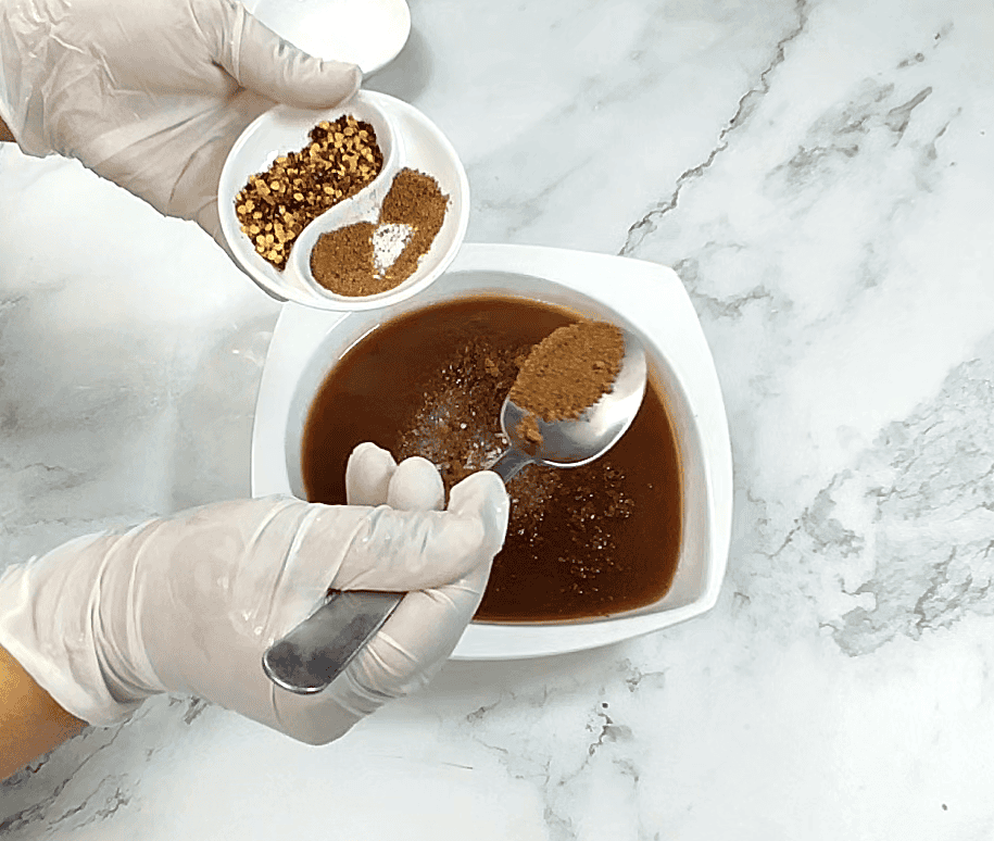 Tamarind chutney being prepared with spices in white bowl, ingredients displayed on marble counter.