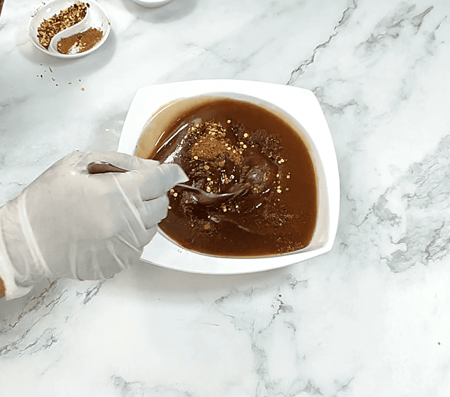 Hand stirring tamarind chutney in white bowl with spice garnish on marble surface.