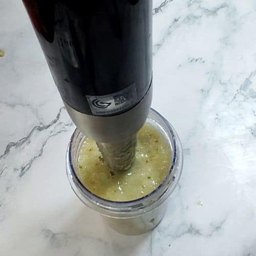 Immersion blender blending yellow-green curry paste in clear container on marble countertop.