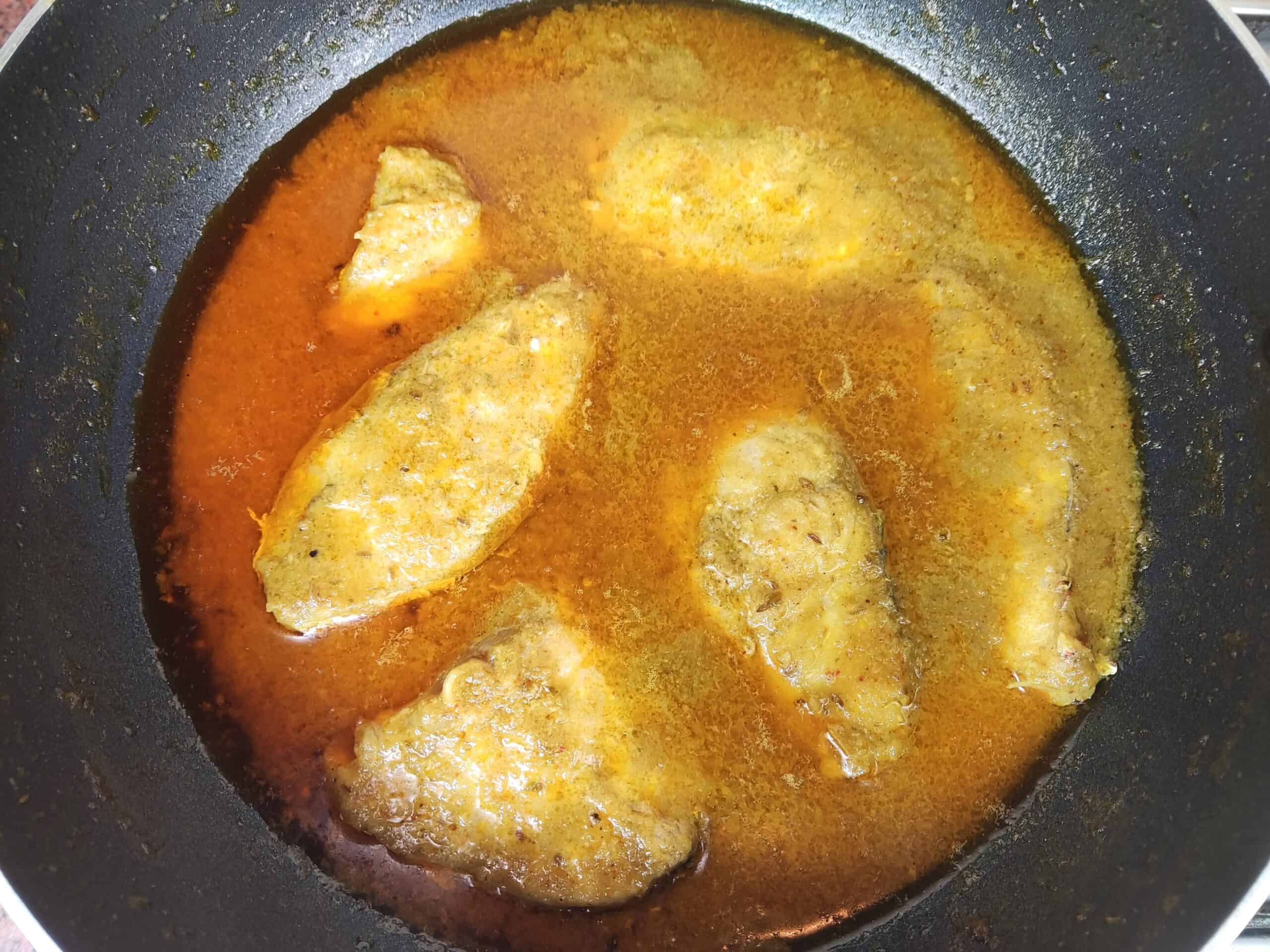 Fish curry simmering in a dark pan with golden-brown fish pieces in spiced sauce.