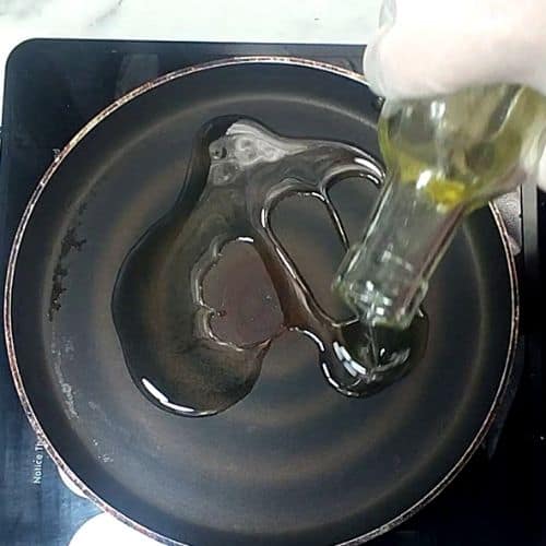 Oil being poured into a dark pan containing raw shami kebab patties for cooking.