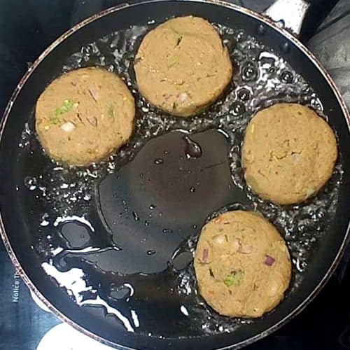Four golden-brown shami kebabs frying in oil in a dark pan with fresh herbs visible.