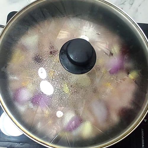 Shami kebab ingredients cooking in covered pot with purple and yellow vegetables in broth.