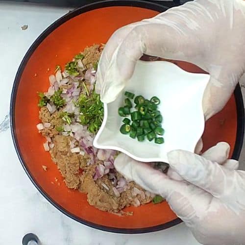 Chef adding sliced green onions to raw shami kebab mixture in orange bowl during preparation.