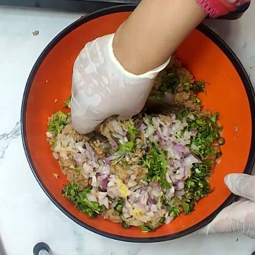 Hand mixing minced meat with herbs and onions in orange bowl for Shami Kebab preparation.