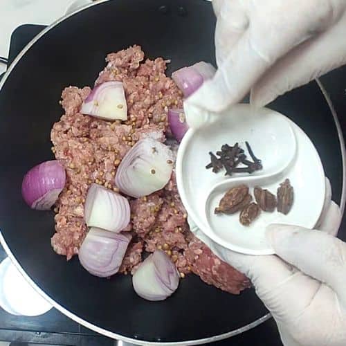 Shami kebab cooking in black pan with onions, cloves, and cardamom spices in small white dishes.