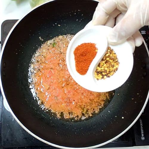 Hand holding white plate with red chili powder and cumin above wok of golden daal mash.