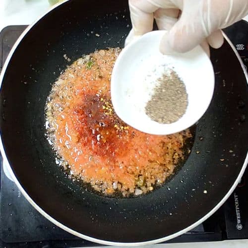 Hand pouring seeds into wok with golden-brown daal mash, Pakistani cooking process.