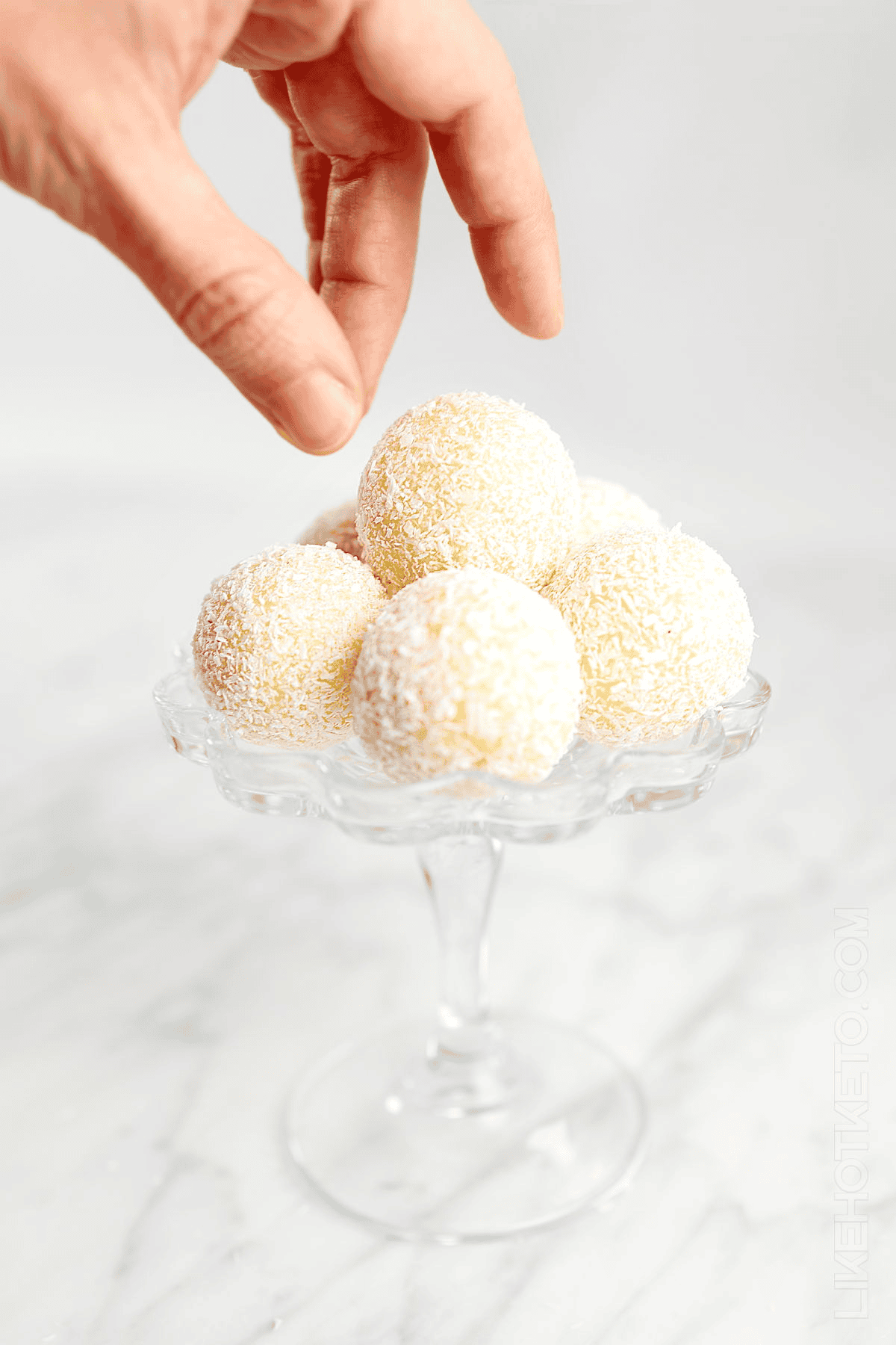 Hand holding coconut ball candy above glass dish with three more golden-coated spheres.