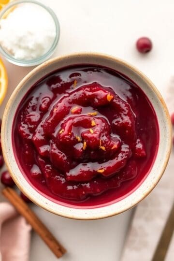 Homemade cranberry sauce in ceramic bowl with orange zest garnish and fresh cranberries nearby.