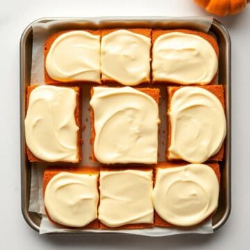 Nine pumpkin bars with cream cheese frosting arranged in a baking pan lined with parchment paper.
