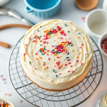 Sugar cookie cheesecake with cream frosting and colorful sprinkles on wire cooling rack.