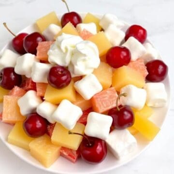 Cherry salad with cubed cheese, fresh red cherries, grapefruit segments, and marshmallows on white plate.