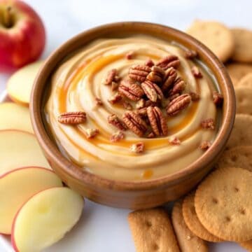 Pecan pie dip in wooden bowl with caramel drizzle, pecans, surrounded by apple slices and crackers.
