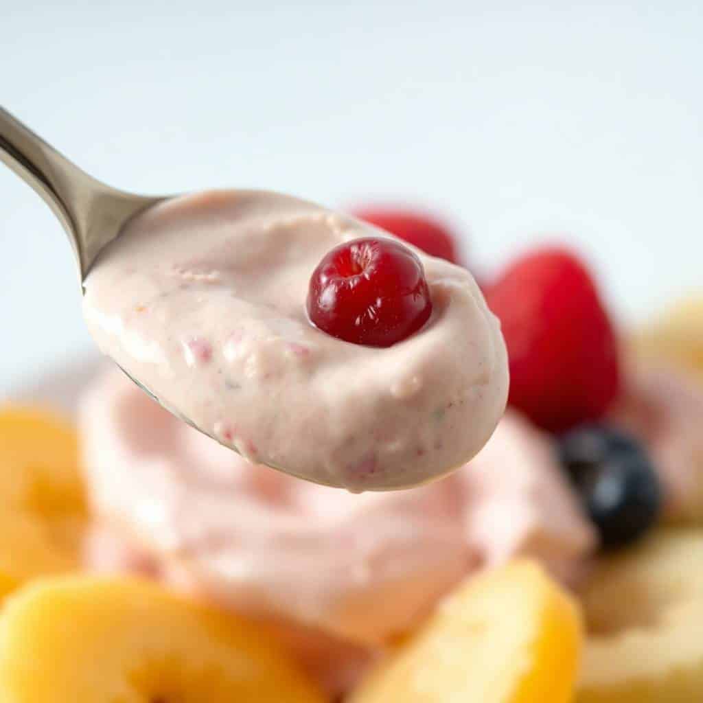 Spoon of cream cheese fruit dip topped with red cherry, surrounded by fresh berries and fruit.