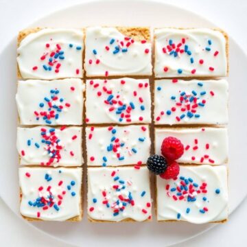 Patriotic sugar cookie bars with white frosting, red and blue sprinkles, arranged on white plate with fresh raspberries.