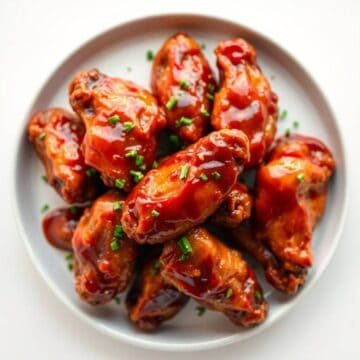 Glazed chicken wings with red sauce and green onion garnish on a white plate.