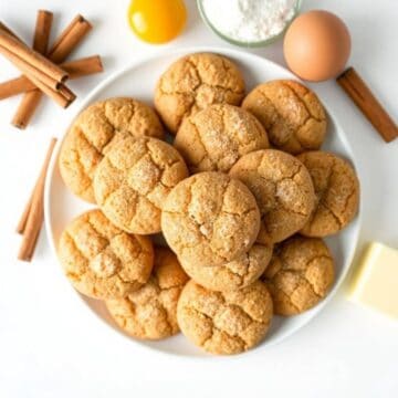 Freshly baked snickerdoodle cookies on a white plate with cinnamon sticks and baking ingredients.