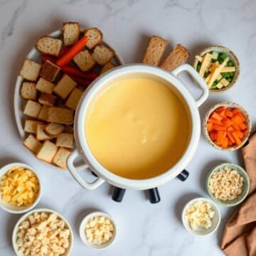 Cheese fondue in a white pot with assorted bread cubes, vegetables, and toppings for dipping.